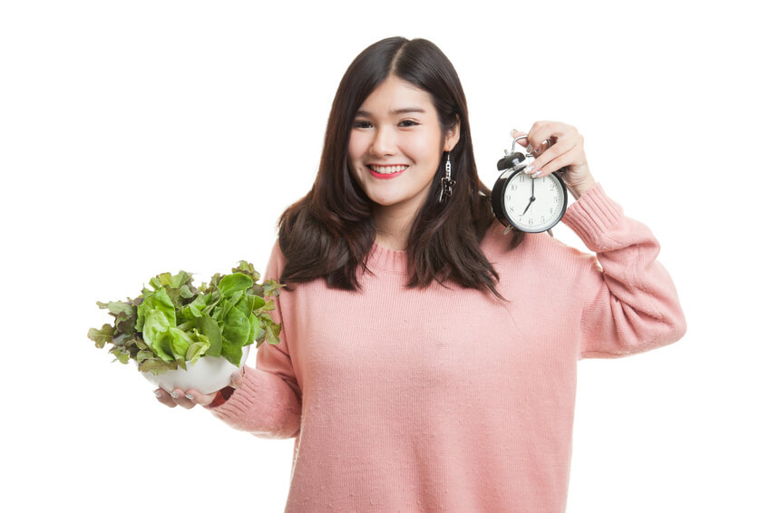 Young Asian woman with clock and salad isolated on white background